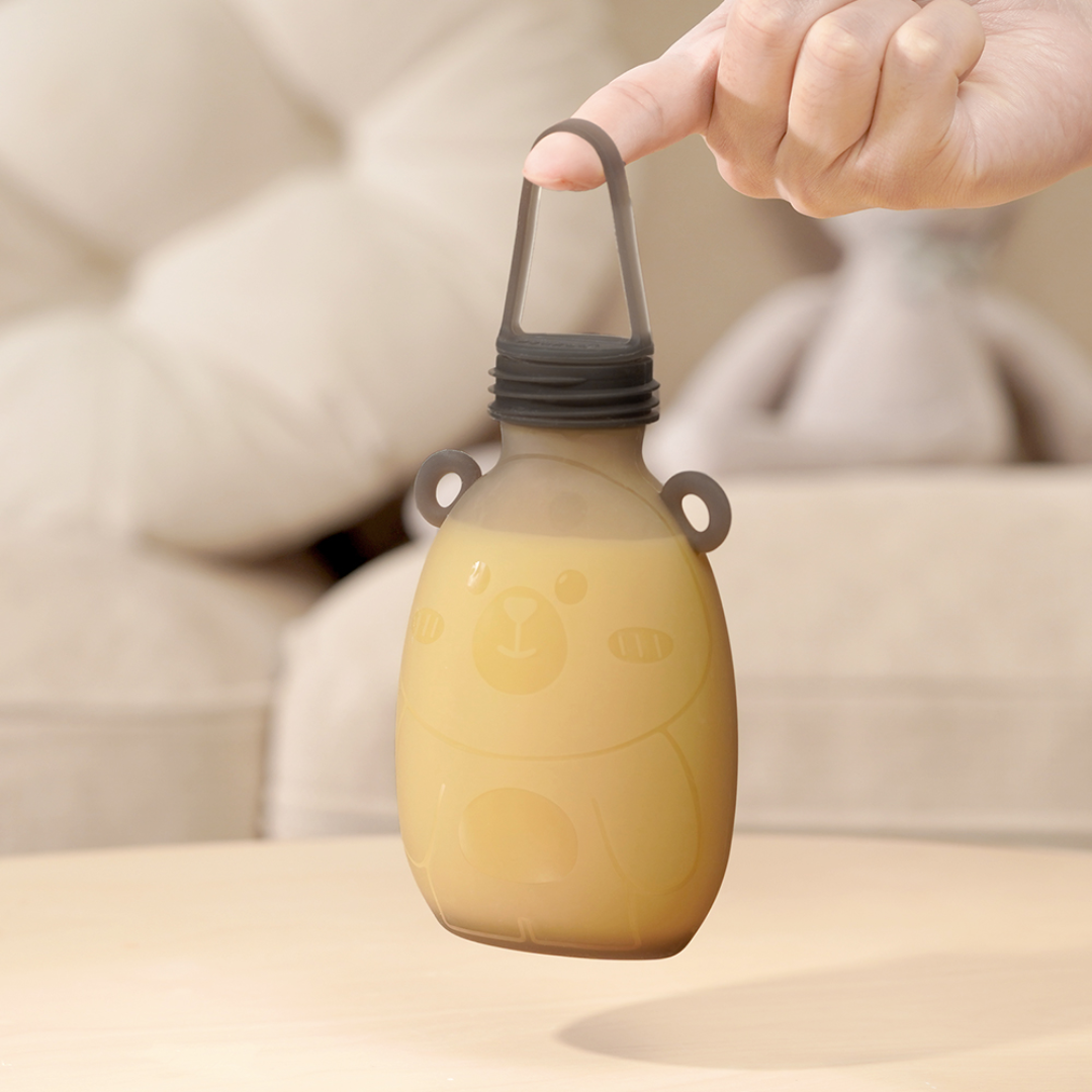 Haakaa Happii Bear Milk Storage Bag, filled with expressed milk and being held by one finger by its carry strap