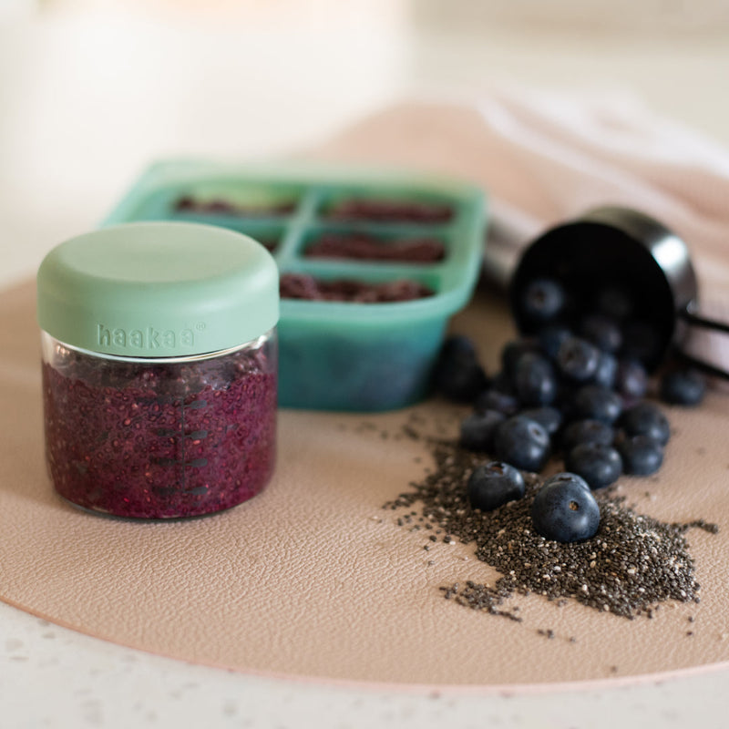 A jar of purple pudding in front of a freezer tray, with a measuring cup of berries and chia seeds spilling out beside them.